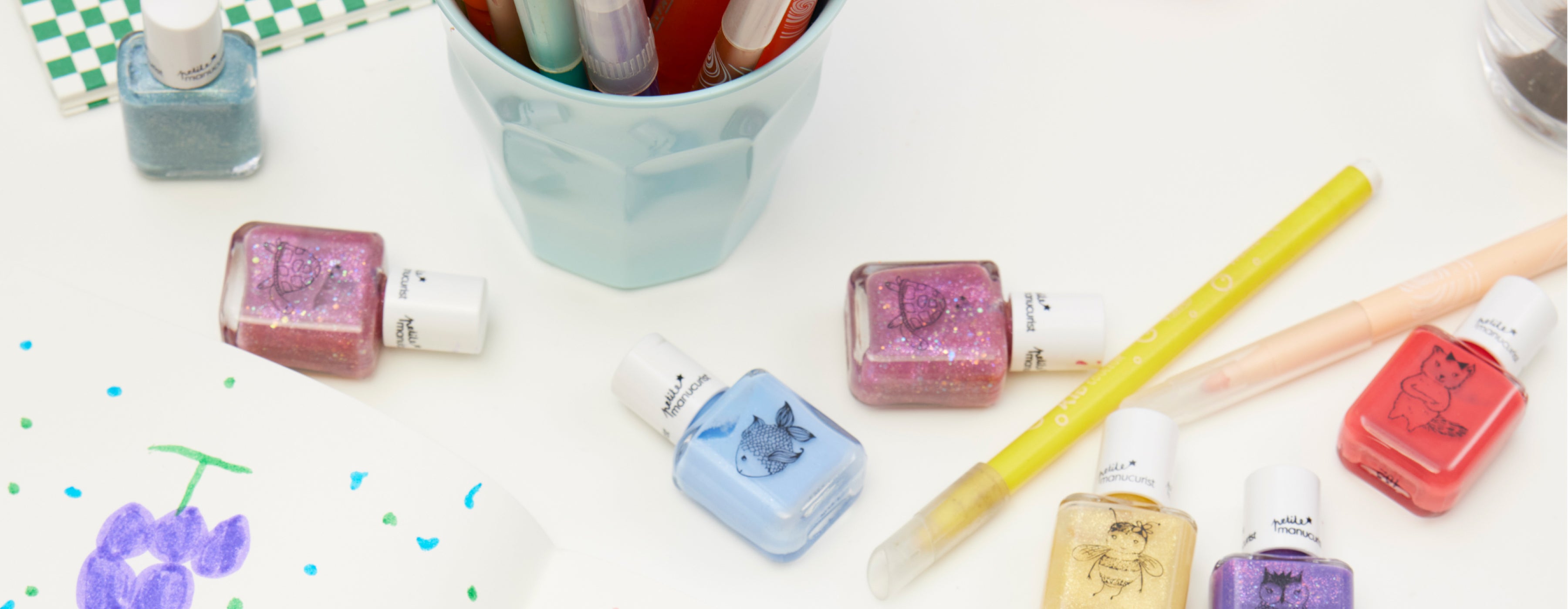 Bottles of colorful nail polish with animal designs, a light blue cup holding markers, two pens, and a drawing on white paper are arranged on a white surface.