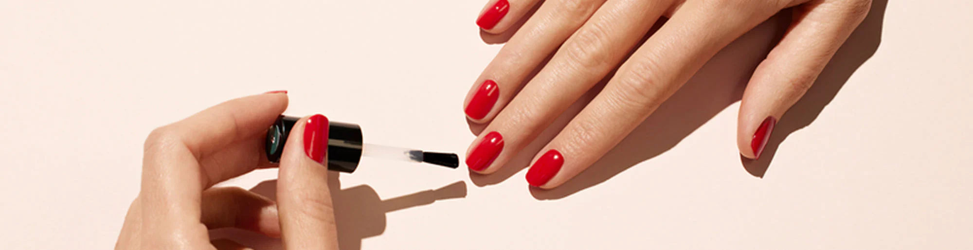 A hand with neatly manicured red nails is being painted with a clear top coat by another hand holding a nail polish brush. The background is a soft, light color.