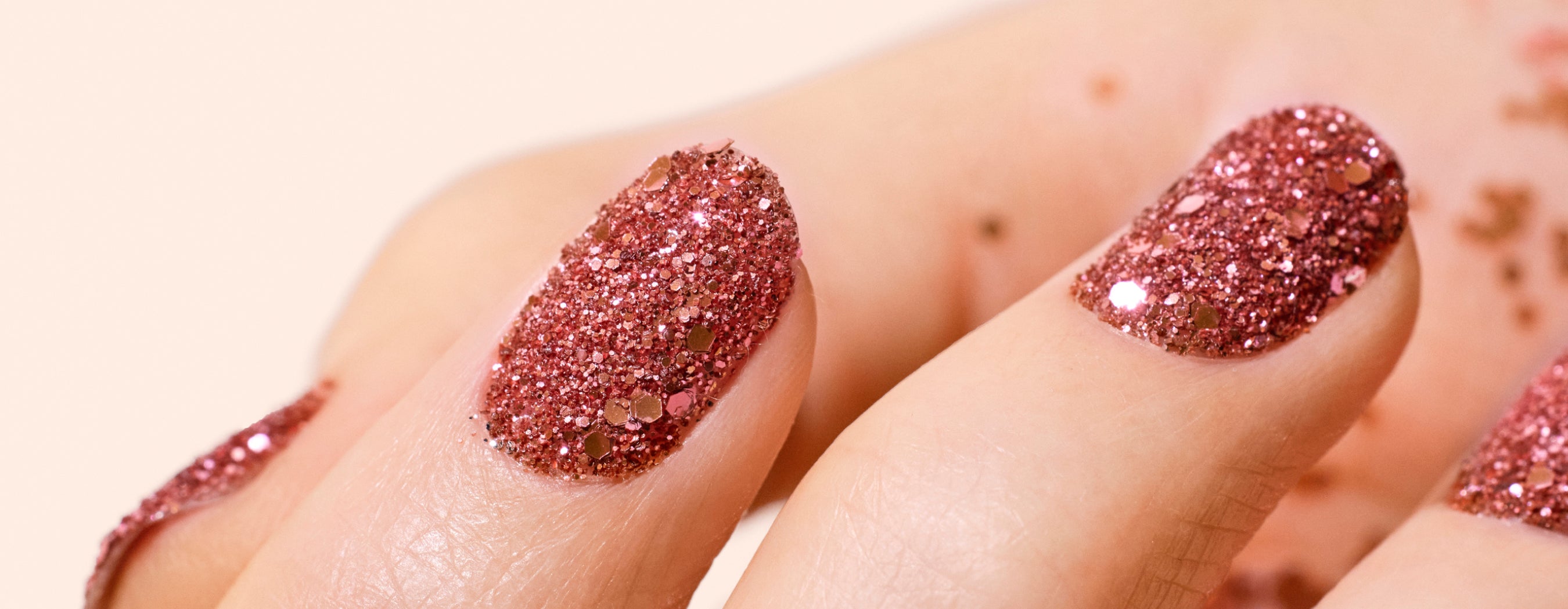 Close-up of three fingers with nails painted in sparkly, pink glitter nail polish. The background is neutral, and some glitter is visible on the skin near the nails.