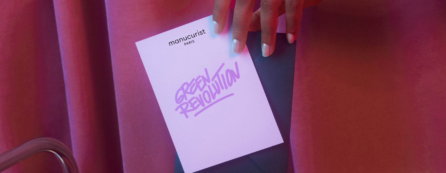 A hand with pastel blue and white nails holds a card reading manucurist paris GREEN REVOLUTION against a pink and purple background.