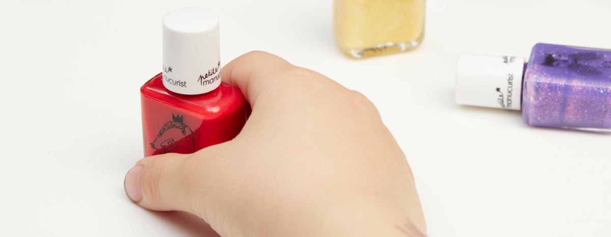 A childs hand holds a red nail polish bottle, with gold and purple nail polish bottles nearby on a white surface.