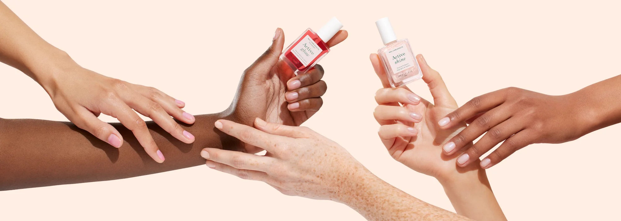 Four hands of different skin tones hold and touch each other, with two of them holding nail polish bottles—one red, one pink—against a light beige background.