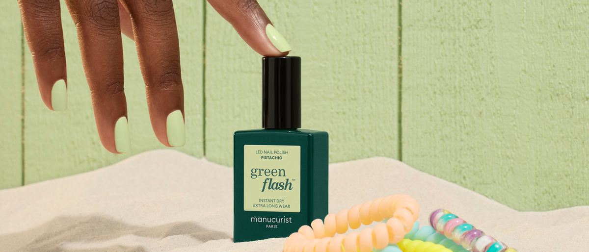 A hand with pale green nail polish reaches for a green flash polish bottle on sand, near pastel-colored bracelets, with a light green wooden background.