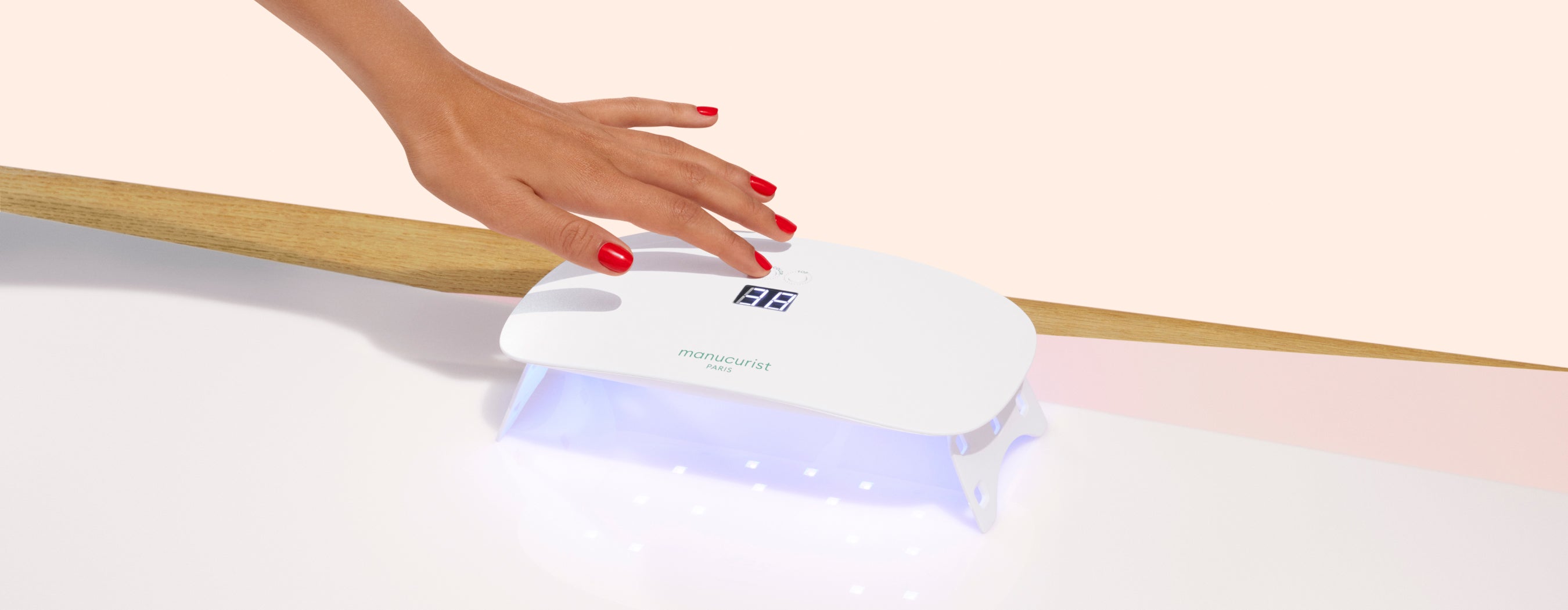 A hand with red painted nails uses a white LED nail lamp with a digital display, positioned on a white and light pink surface.
