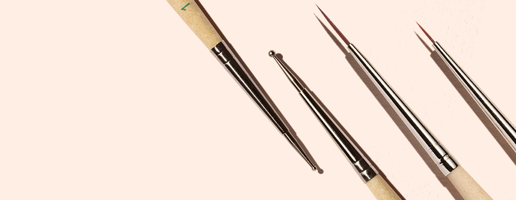 Four nail art brushes with wooden handles and metal tips are arranged diagonally on a light beige background.