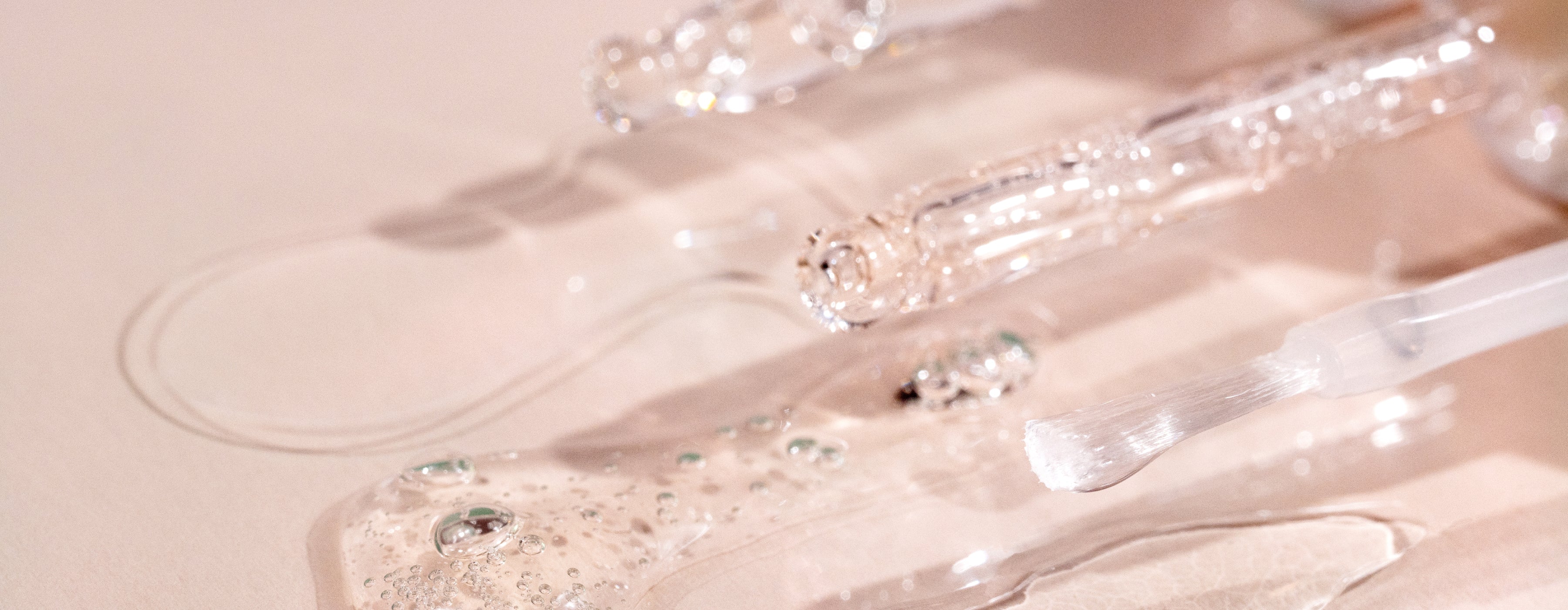 Close-up of clear glass droppers and a brush applicator with transparent gel serum and bubbles spilled on a light, reflective surface. The scene is softly lit with a pinkish hue.