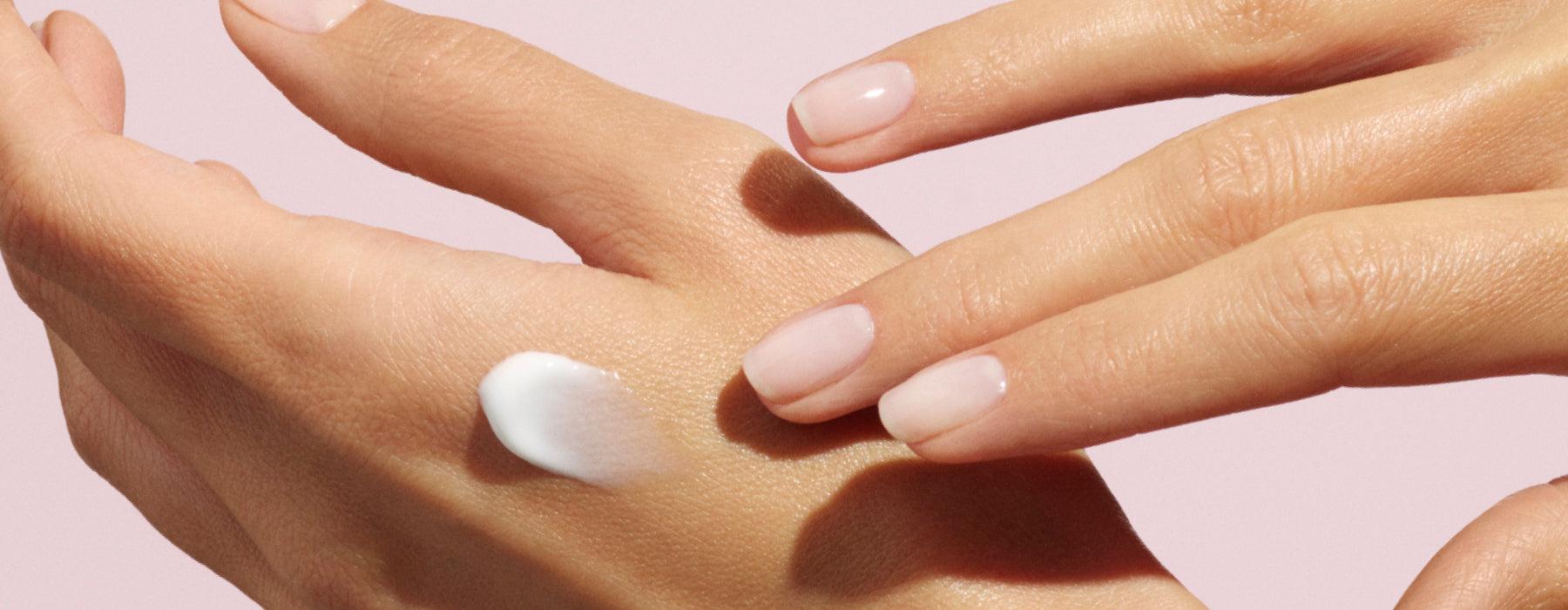 A close-up of a hand applying a dab of white lotion onto the back of another hand, with natural, neatly manicured nails against a pale pink background.