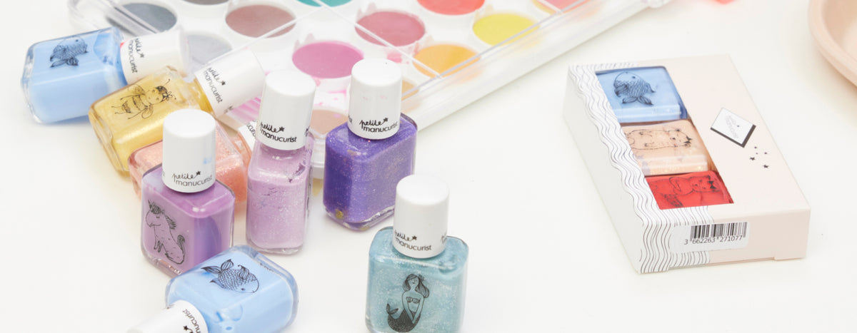 Nail polishes in pastel colors, a watercolor paint palette, and a small box with two pastel eyeshadows are arranged on a white surface.