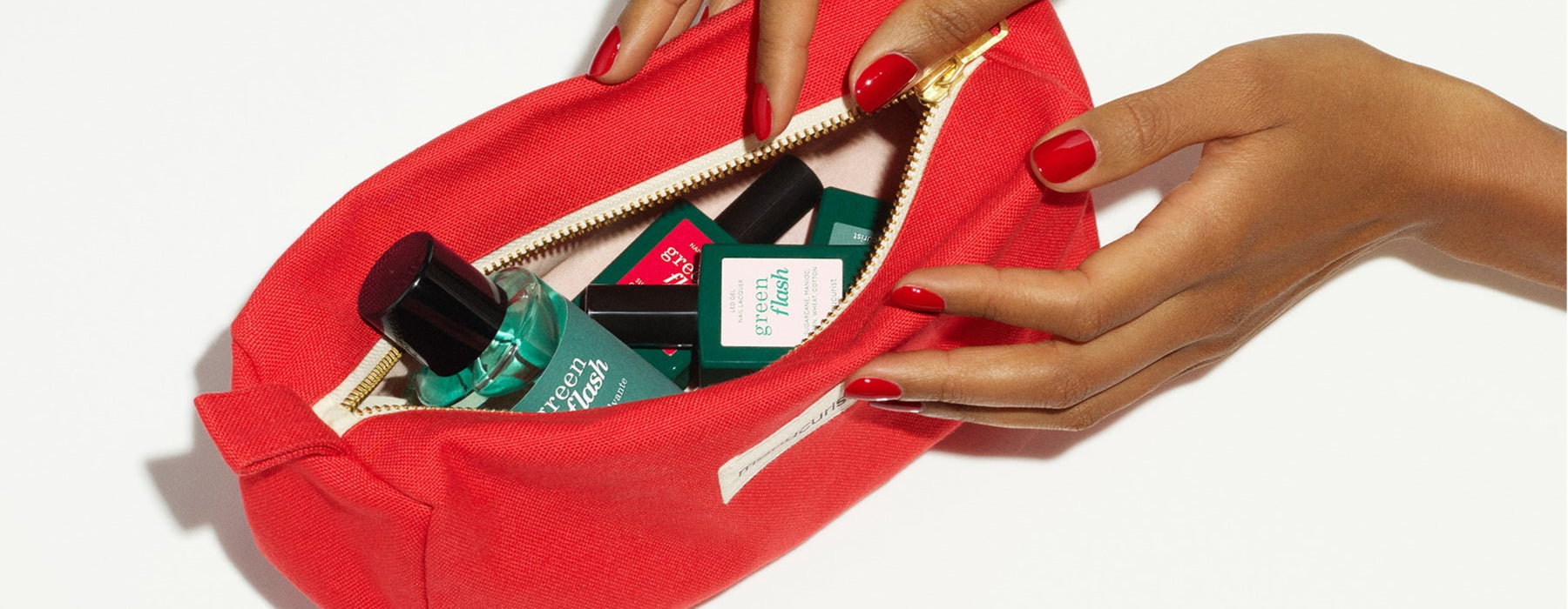 A person with red nail polish opens a red zippered pouch filled with green and black bottles of beauty products, against a white background.