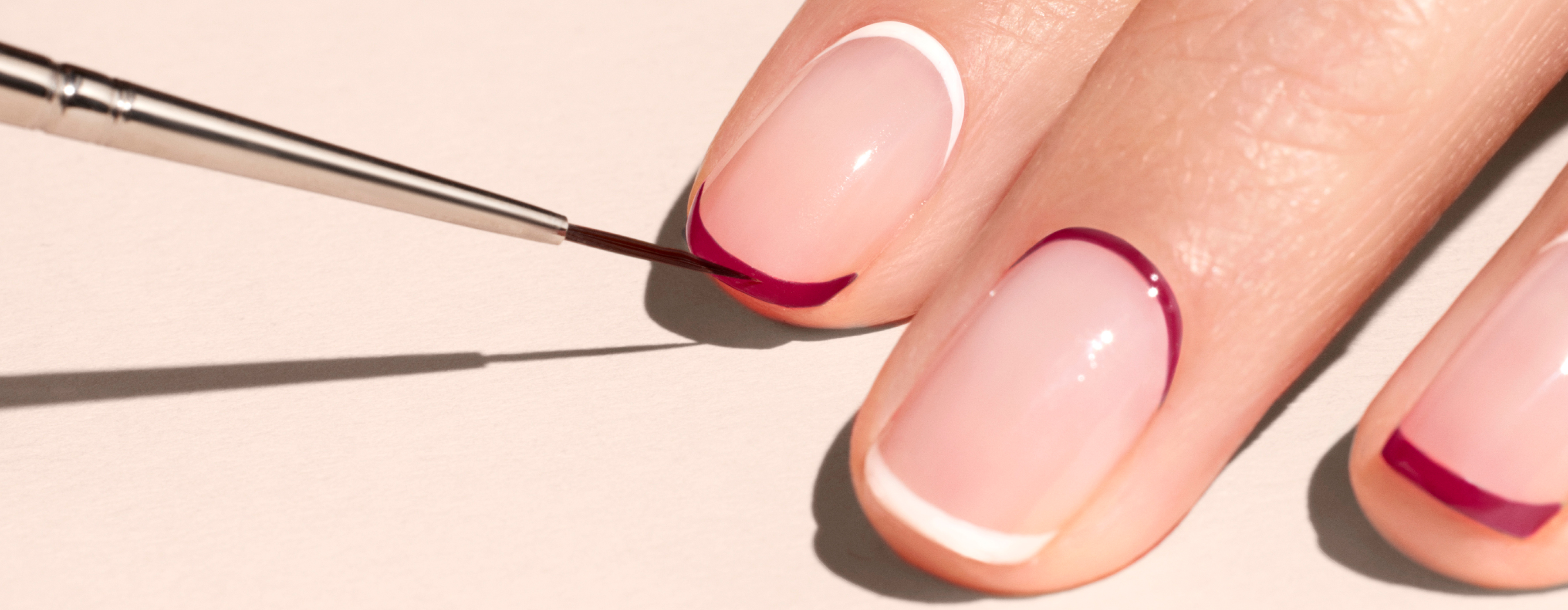 A close-up of a hand with manicured nails, featuring a thin brush painting a dark red French tip on one nail. The other nails have white and dark red French tips.