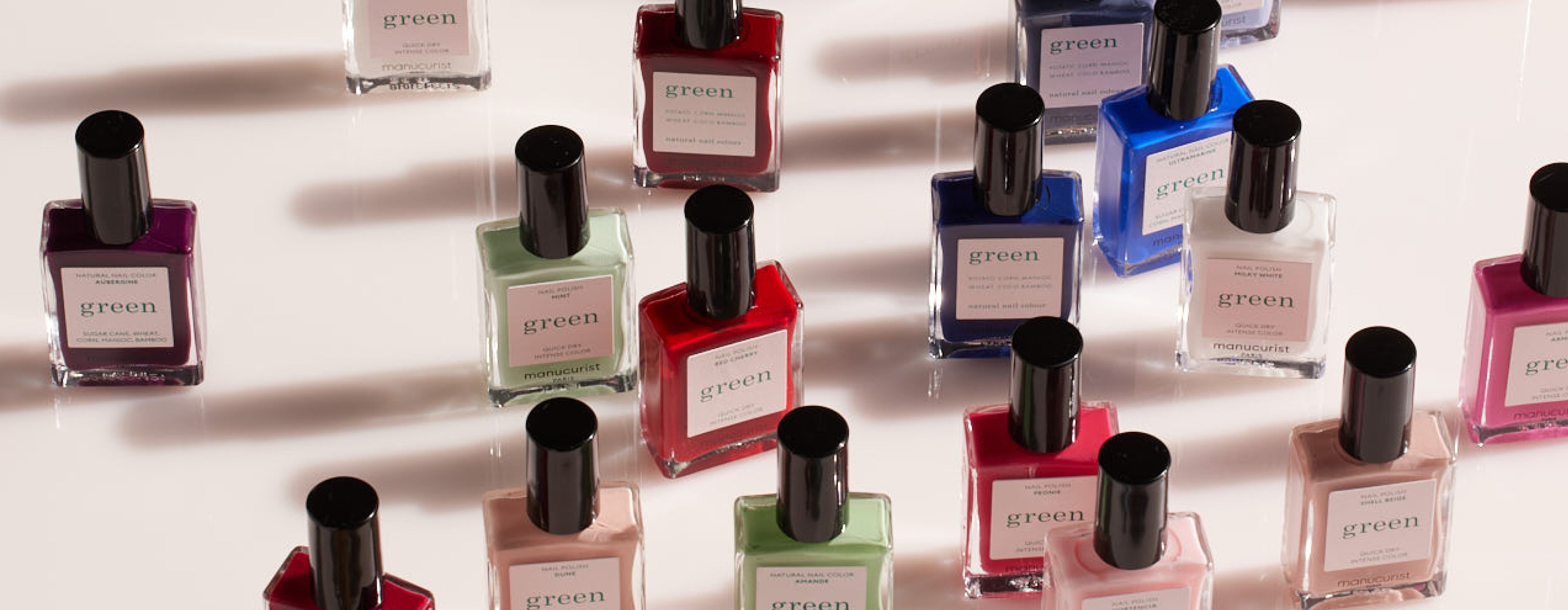 Several bottles of nail polish in various colors, including red, pink, purple, green, and blue, are arranged on a light surface. The bottles are labeled “green” with black caps.