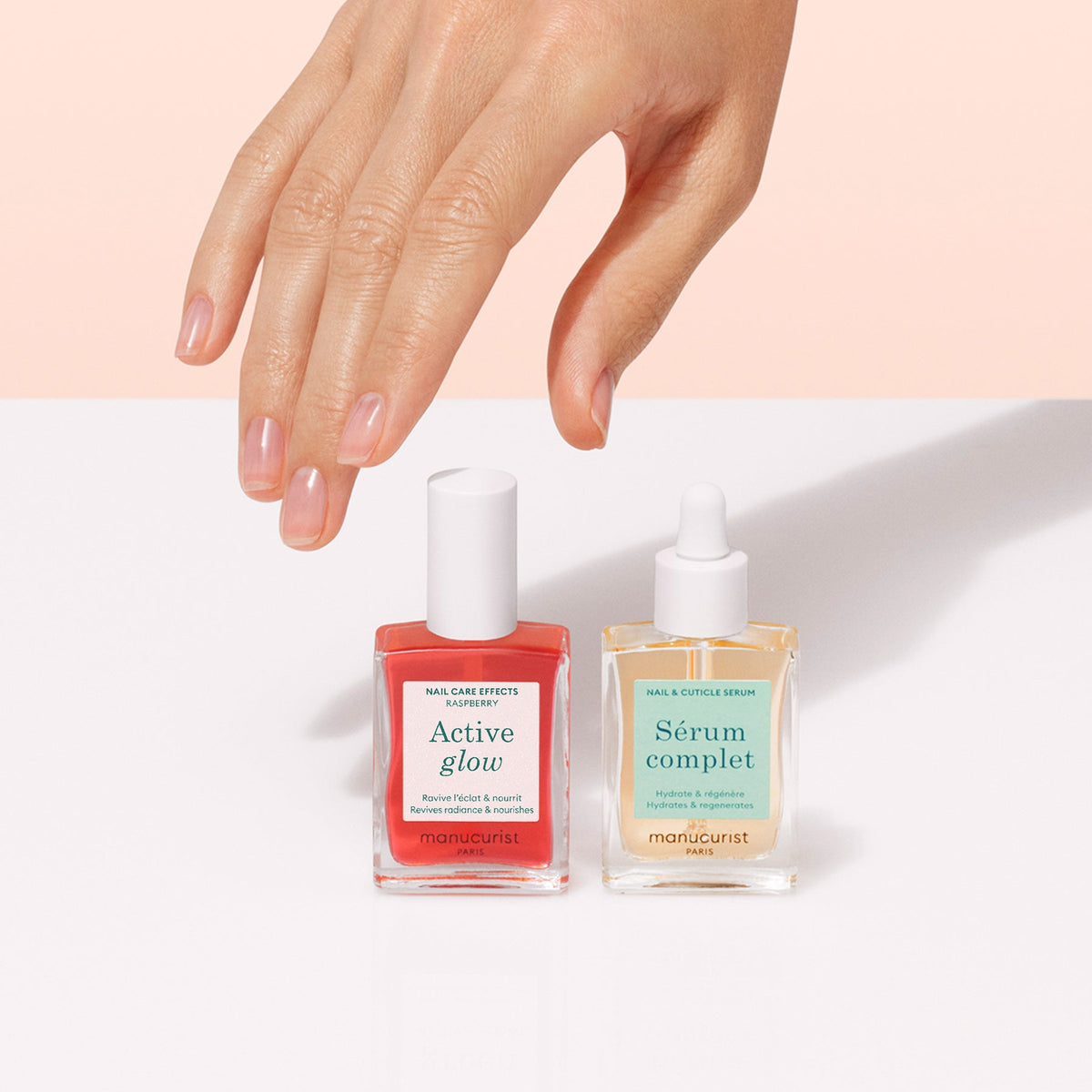 Une main aux ongles soigneusement manucurés est posée au-dessus du Duo Healthy Glow de Manucurist — deux flacons de soin pour les ongles — sur une surface blanche sur fond rose poudré.