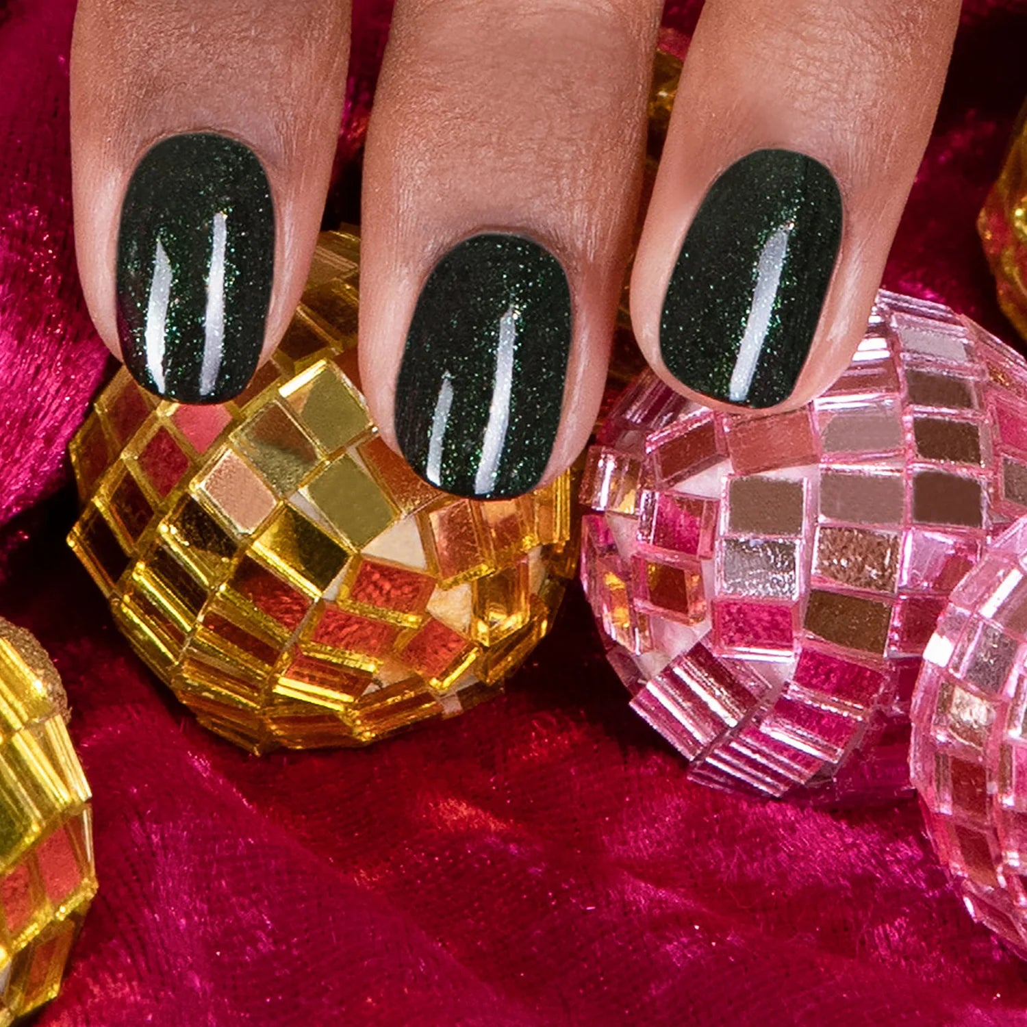 A hand with short, oval-shaped nails painted dark green with subtle glitter rests on red fabric, surrounded by gold and pink mini disco ball ornaments.