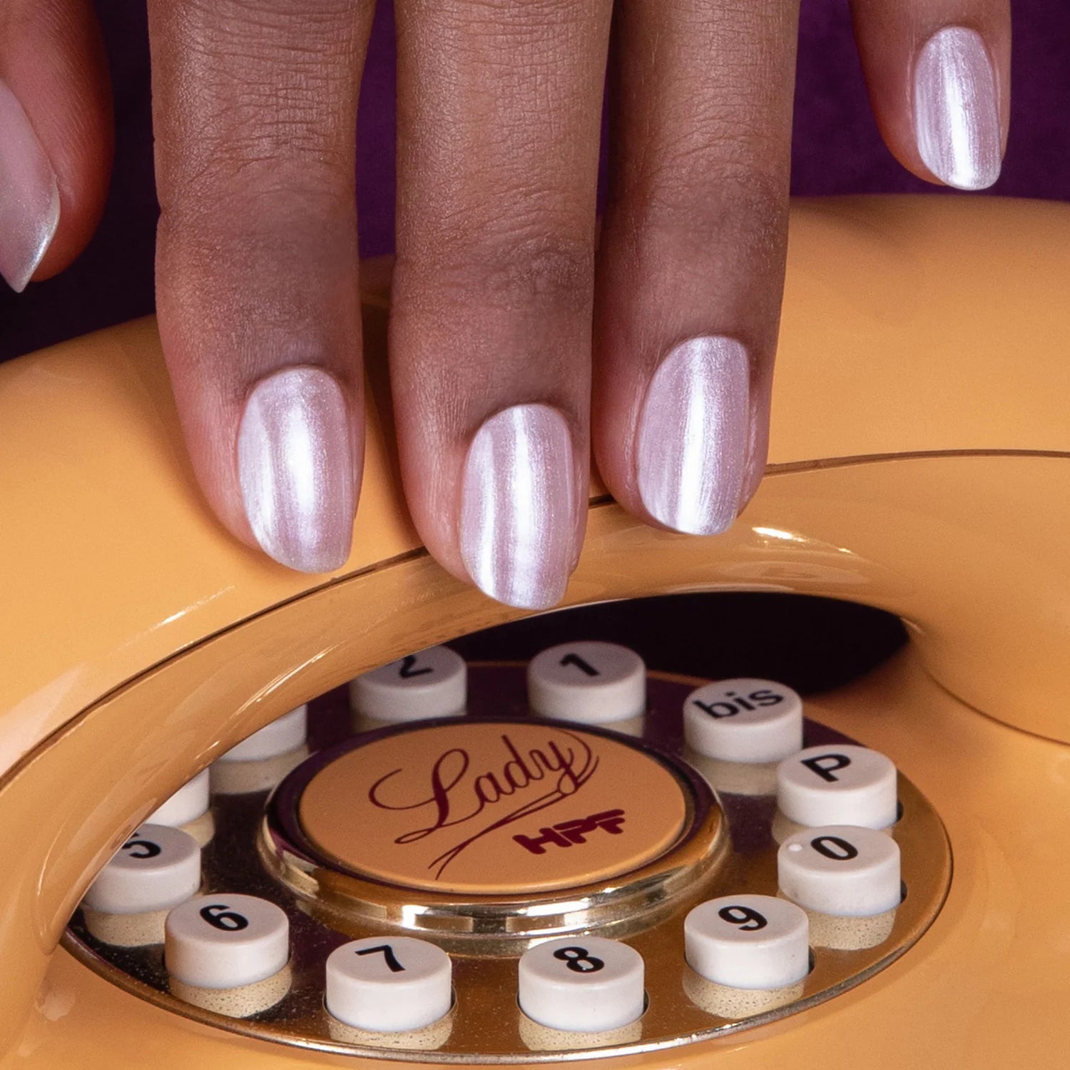 A close-up of a hand with shiny, light pink manicured nails resting on the rotary dial of a vintage yellow telephone labeled Lady HFF.