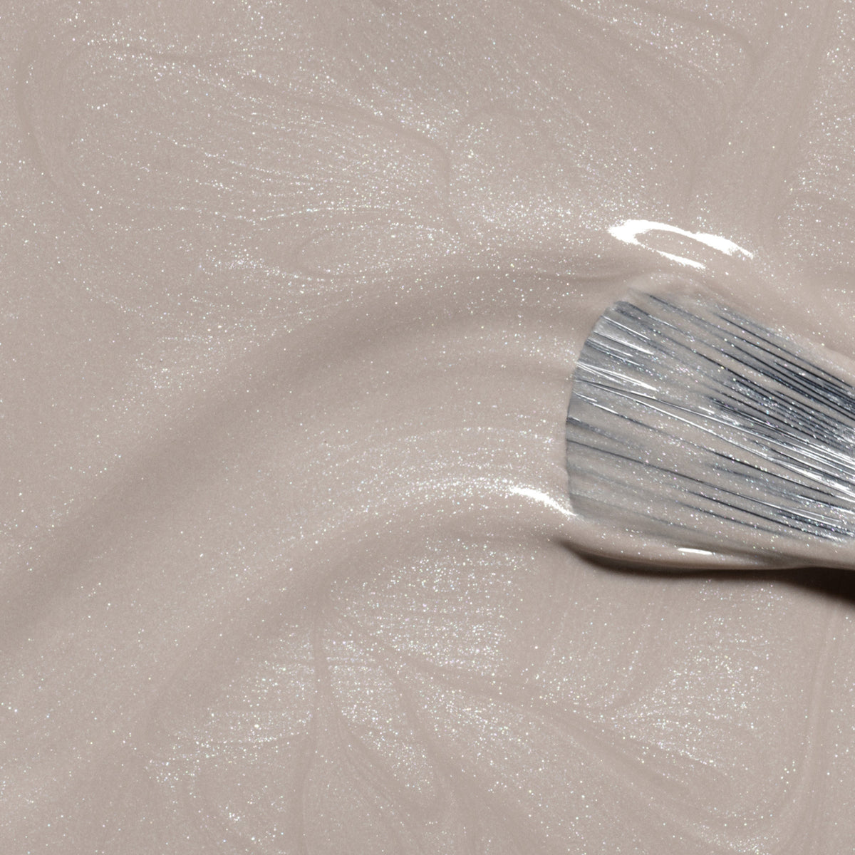A close-up of a brush applying Manucurist Tweed, a shimmery light beige nail polish with pearly undertones, creating gentle swirls in the glossy finish.
