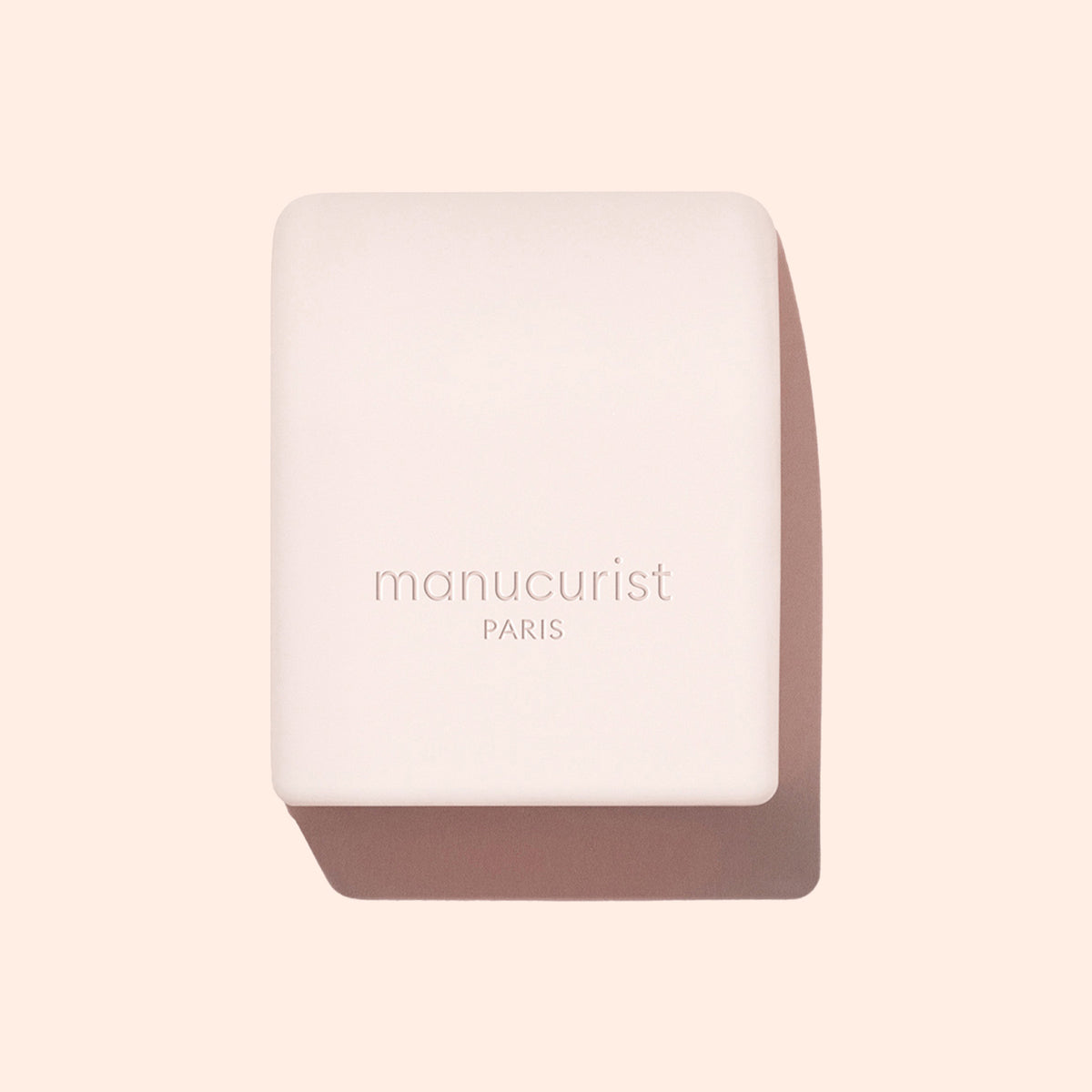 A pale pink rectangular Mani Helper soap bar from Manucurist US, embossed with the logo, rests on a beige background with a soft shadow—ideal for prepping hands before a perfect manicure or nail polish application.