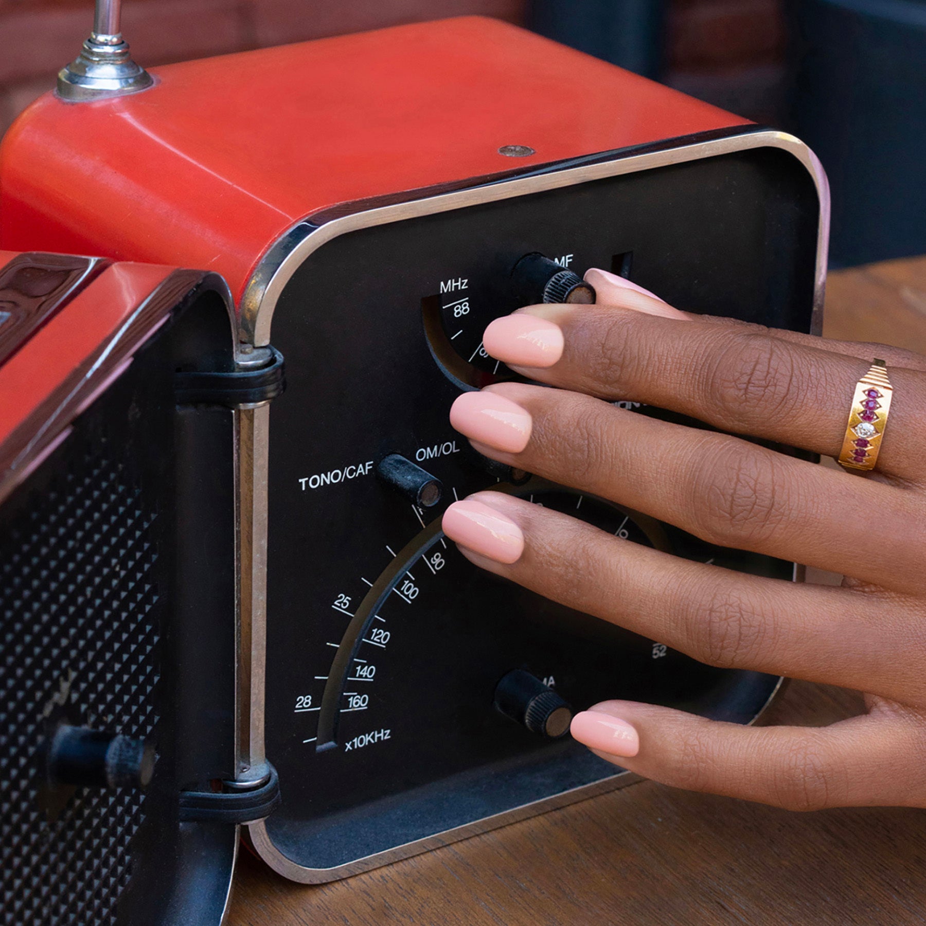 A hand with Manucurist Dune gel polish and a gold ring adjusts the dial of a vintage red radio on a wooden surface.