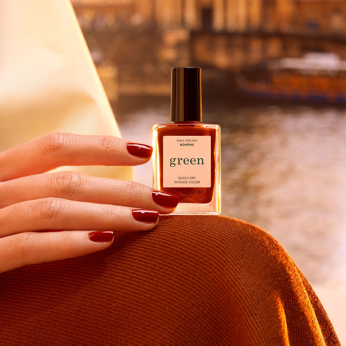 A hand with Montmartre-inspired dark red Bohème polish by Manucurist, featuring brown undertones, holds a green polish bottle against a warm, blurred outdoor backdrop for a cozy, elegant vibe.
