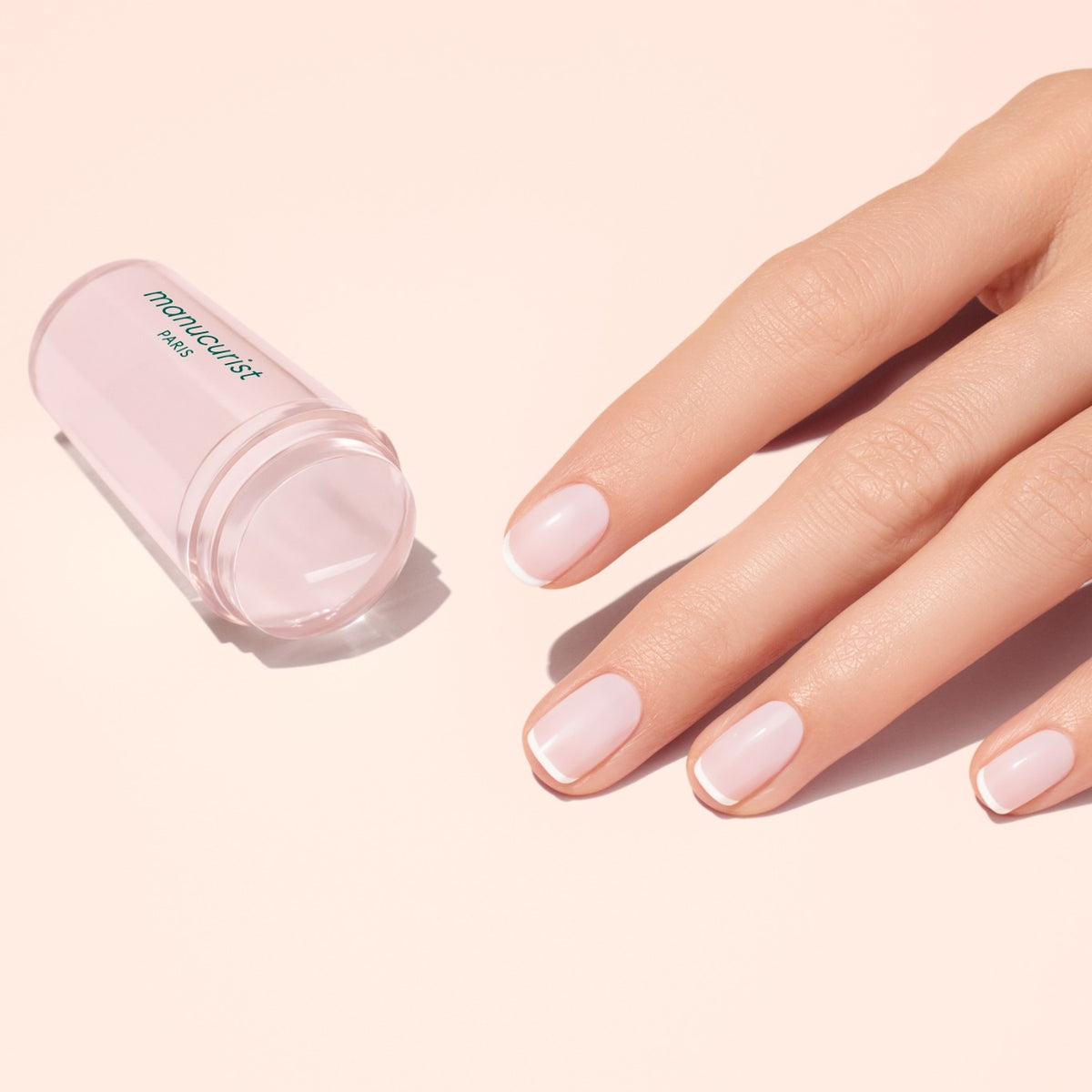 Une main avec une French manucure impeccable repose à côté d’un tampon Mani stamper rose translucide par Manucurist US sur une surface rose clair.