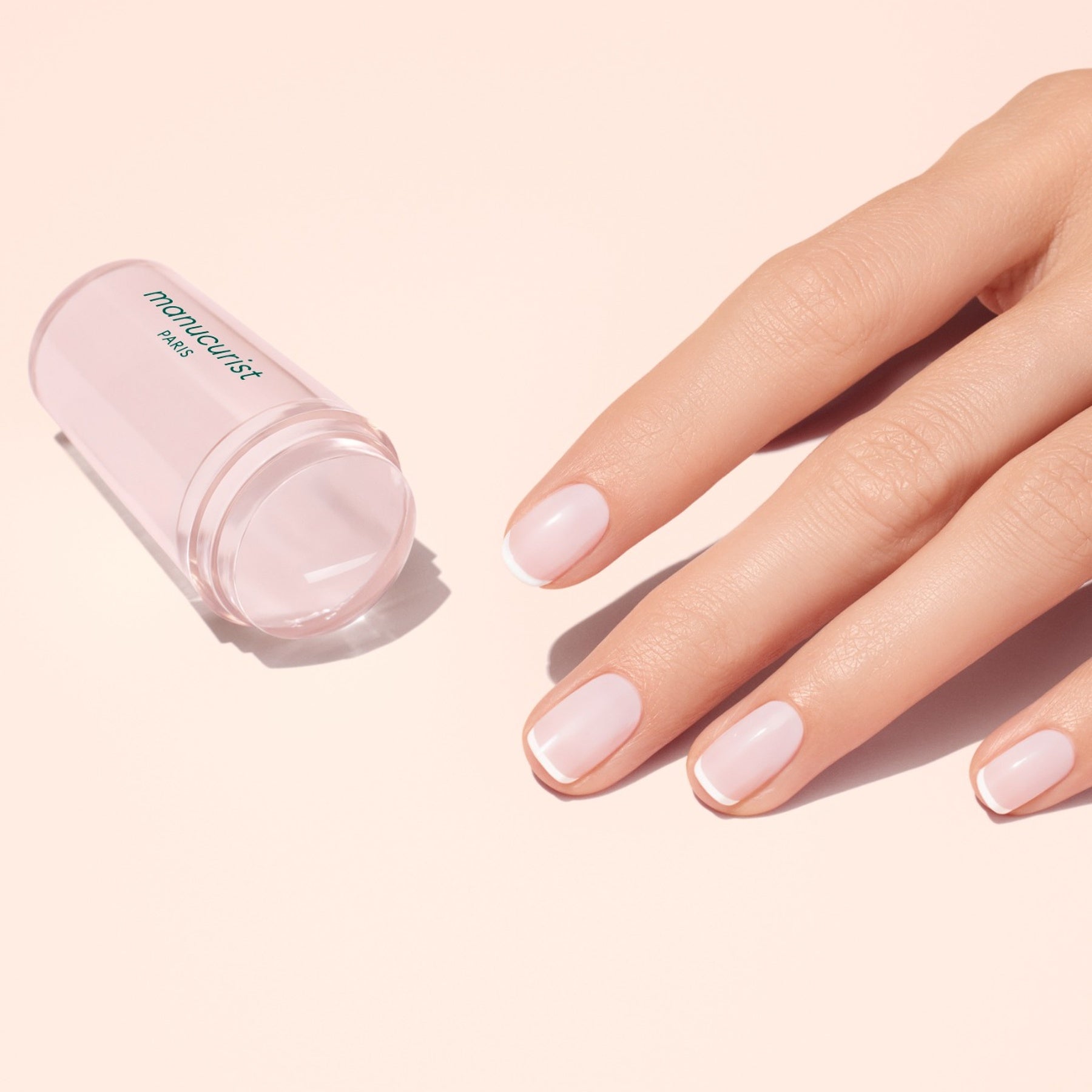 A hand with neatly manicured, shiny pale pink nails featuring elegant French tips rests on a light pink surface beside a transparent container labeled Manucurist French Active™ Kit.