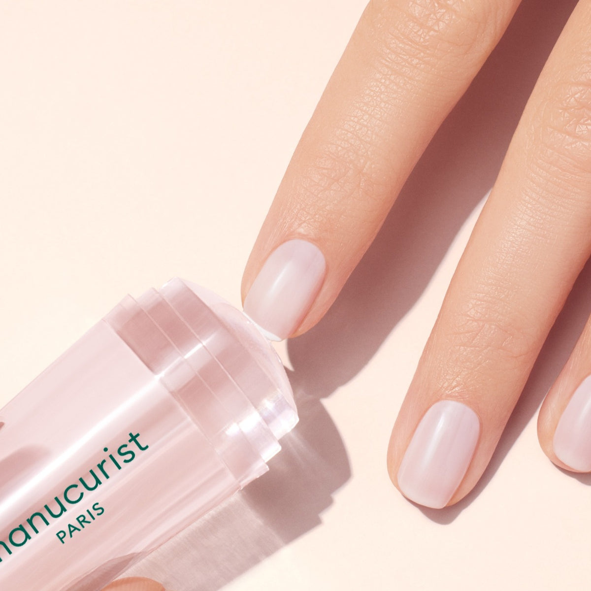 Une main aux ongles soignés et naturels applique le stick stamper transparent Manucurist US Mani, idéal pour un French manucure impeccable ou une base lisse avant le stamping, sur fond clair.