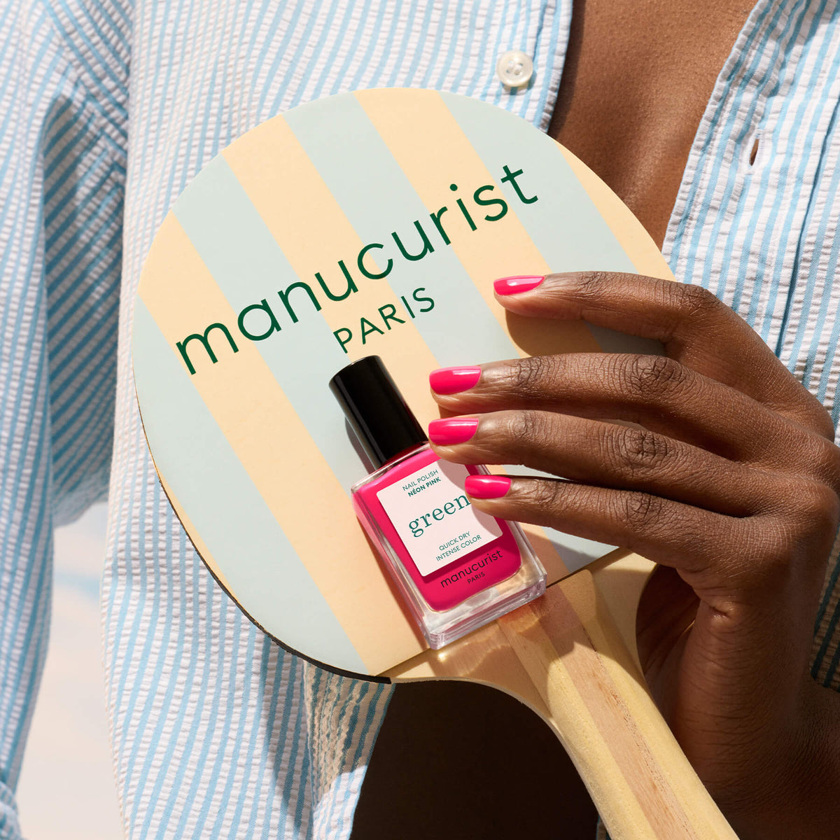A hand with Neon Pink Manucurist polish holds a striped ping pong paddle labeled Manucurist, with a matching semi-matte bottle resting on it. The person wears a blue striped shirt, giving off summer vibes.