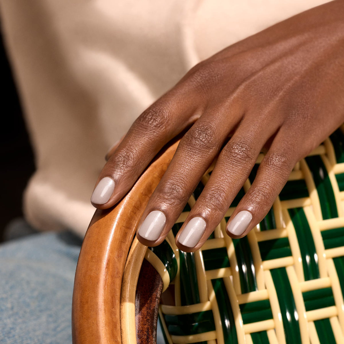 A hand with neatly manicured, light beige nails polished in "Tweed" by Manucurist rests on the curved wooden arm of a woven chair with green and yellow accents. The softly blurred background adds to the elegant look.