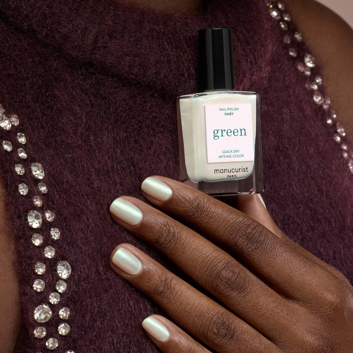 Une main portant un vernis vert clair pailleté métallique tient un flacon assorti étiqueté "Fairy" par Manucurist, sur fond de haut bordeaux orné de strass.