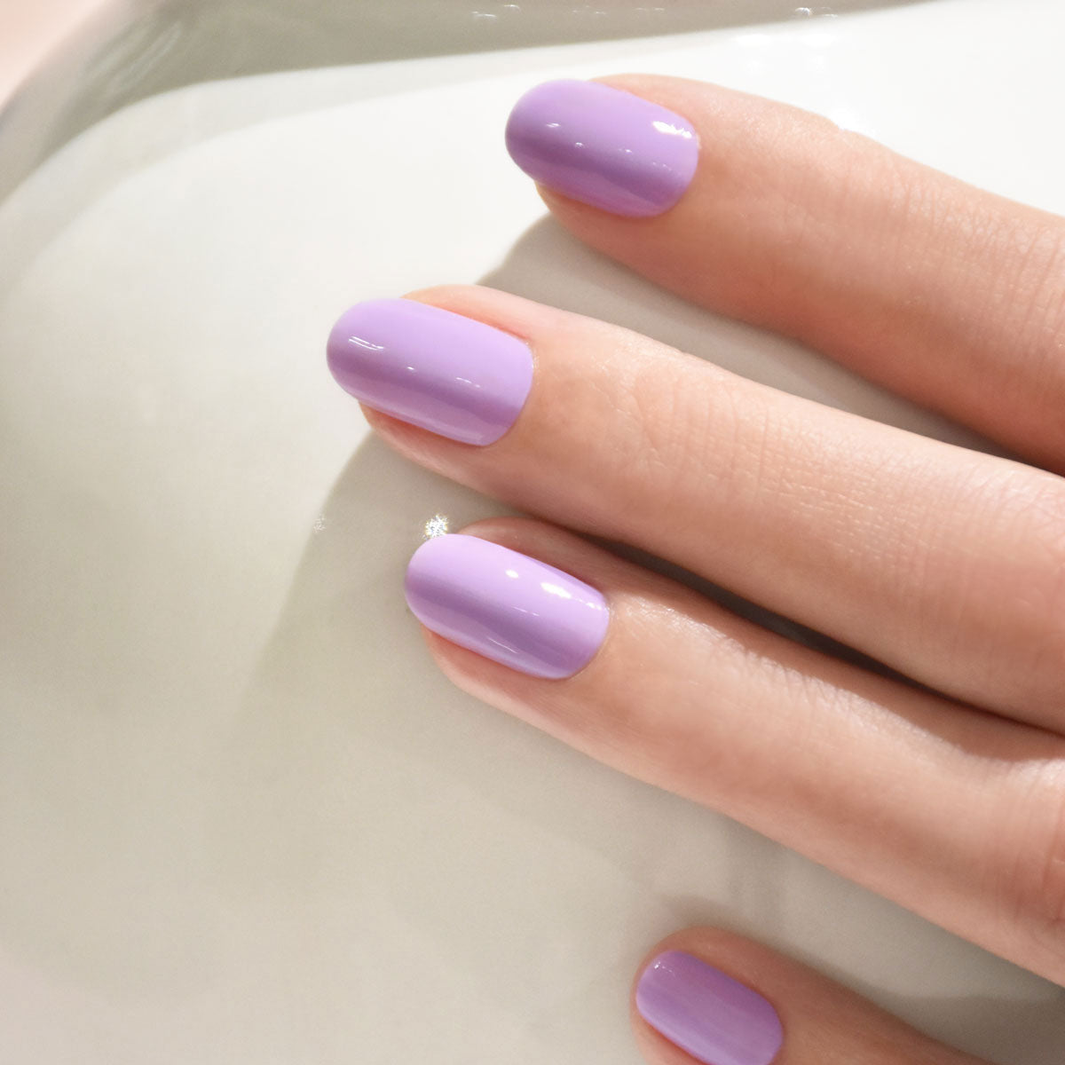 A close-up of a hand with neatly manicured nails painted in a glossy lavender purple color, resting on a smooth, white surface.