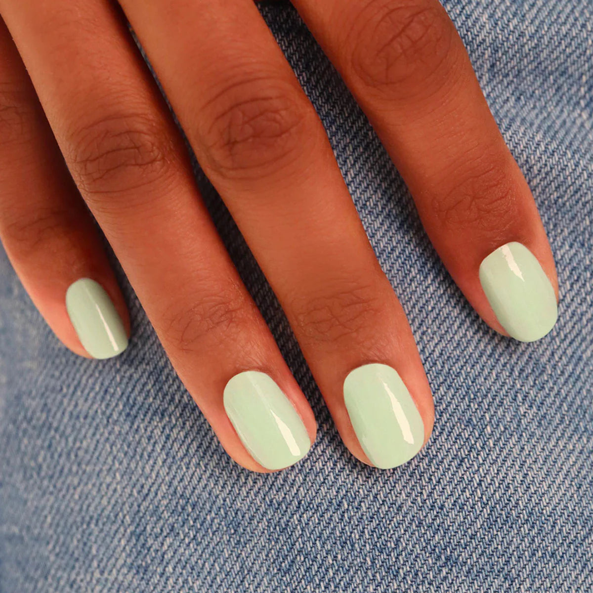 A hand with short, neatly manicured nails painted in a glossy pastel mint green color rests on blue denim fabric.