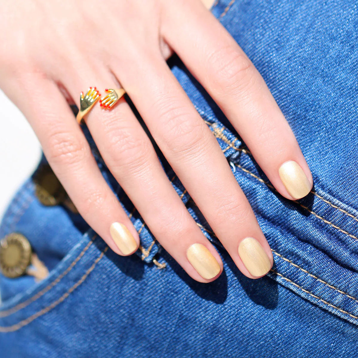 A hand with gold metallic nail polish rests on blue denim jeans. The person wears a gold ring with a unique design on their middle finger.