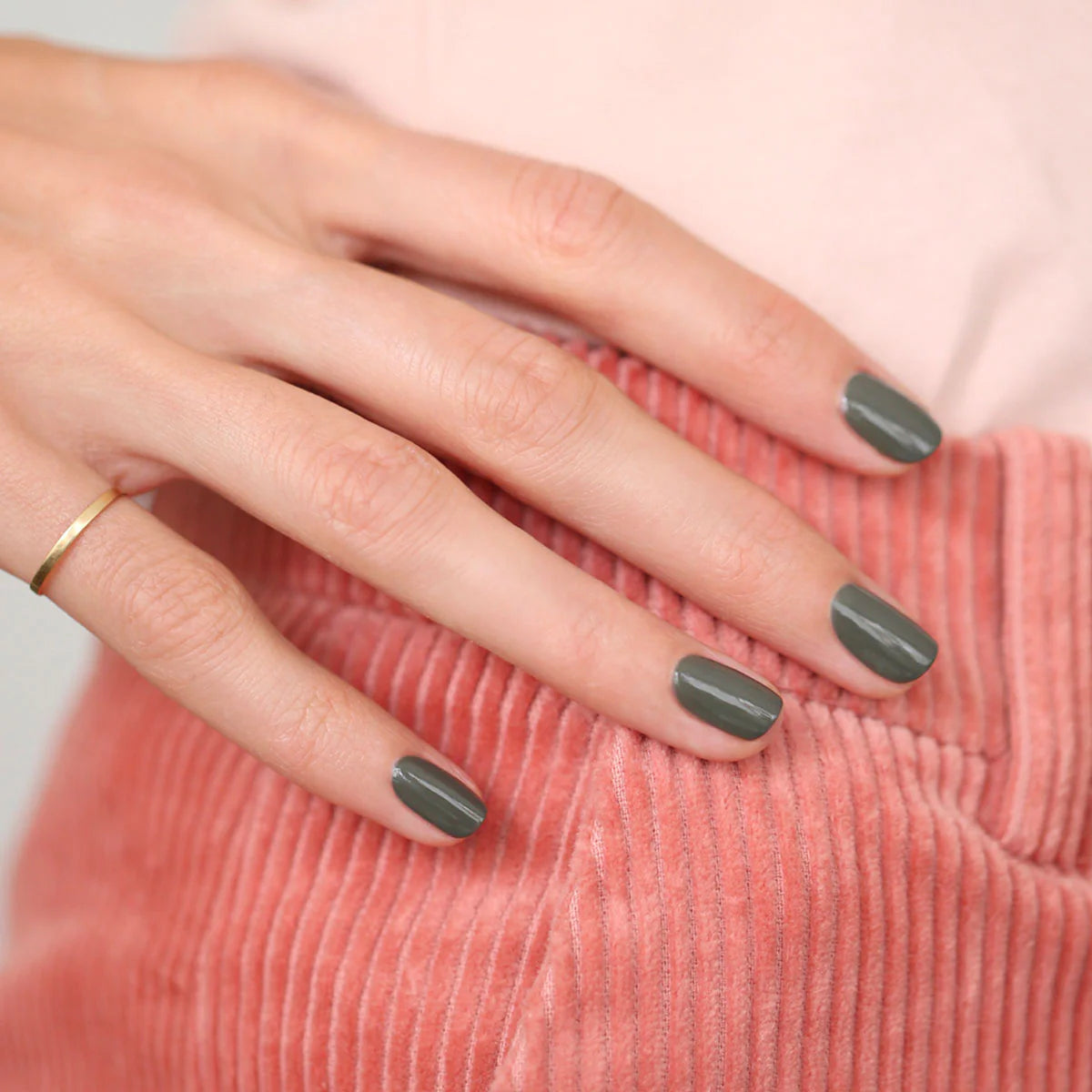 A hand with short, neatly manicured nails painted in a dark gray shade rests on pink corduroy fabric. A simple gold ring is worn on the ring finger.