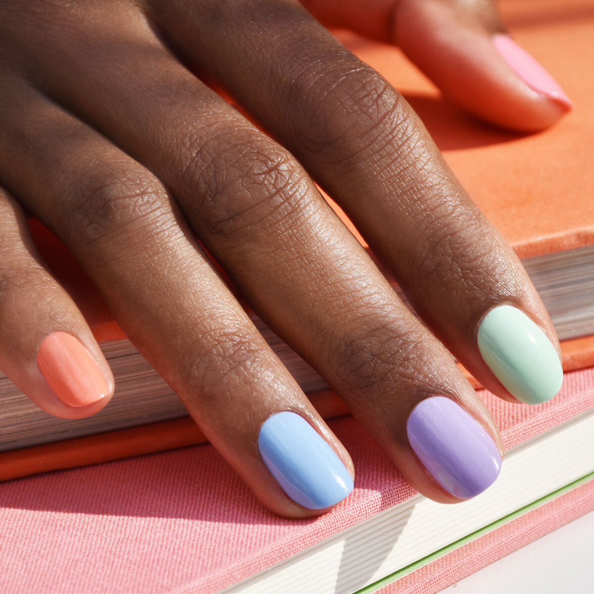 A close-up of a hand with nails painted in pastel colors—peach, blue, purple, and green—resting on stacked books with pink and orange covers.