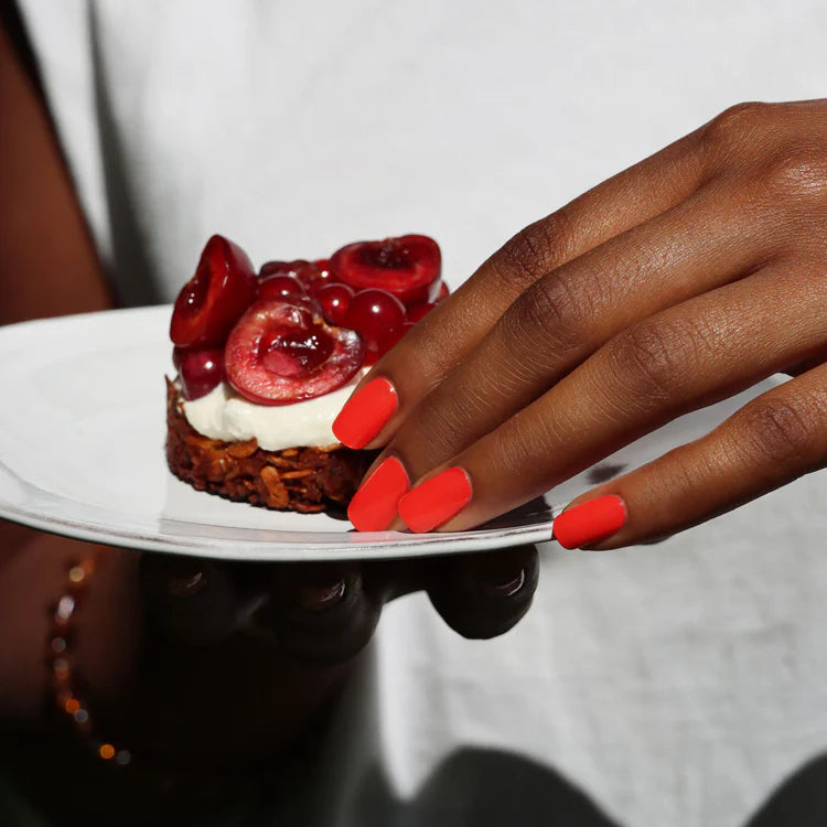A hand with bright orange nail polish holds a white plate with a dessert topped with whipped cream and fresh cherries.