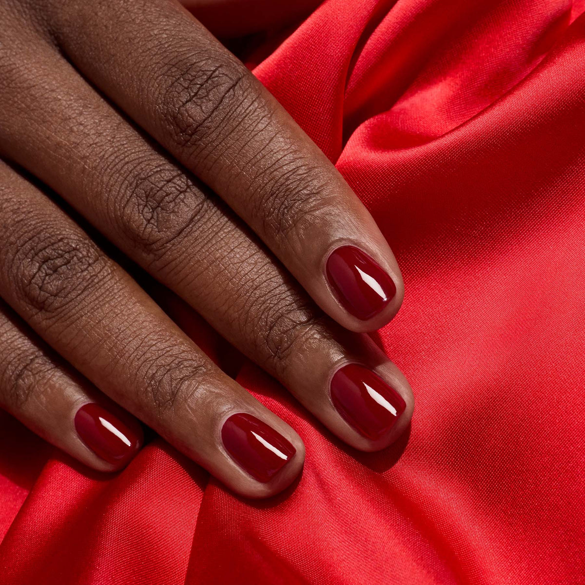 Gros plan sur une main aux ongles soignés vernis en Dark Pansy par Manucurist, posée sur un tissu satiné rouge vif.