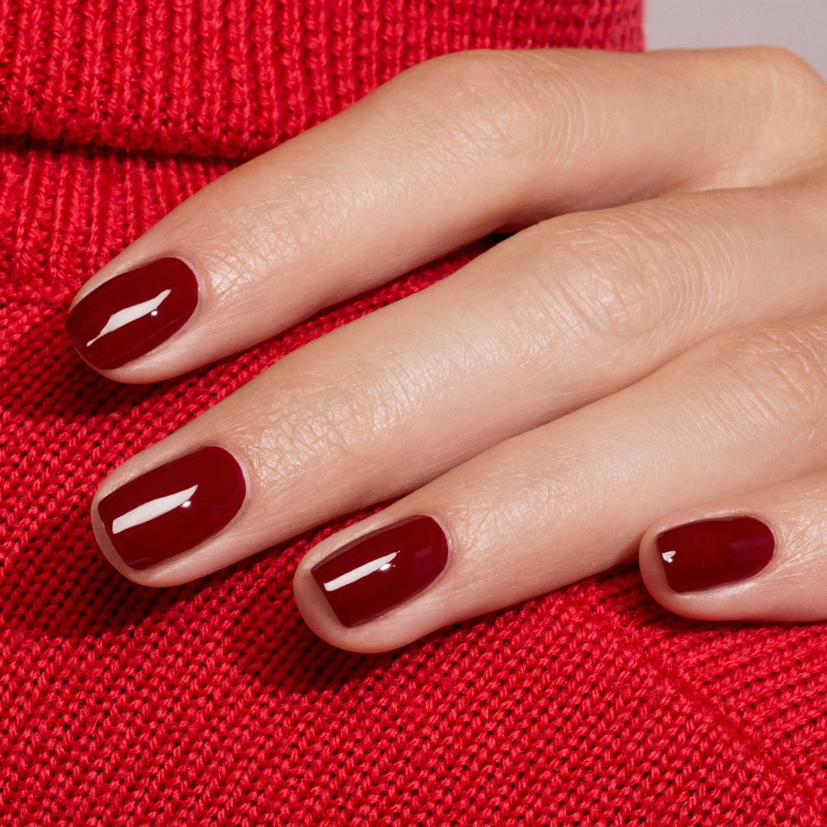 Une main aux ongles impeccablement manucurés, vernis avec la teinte Dark Pansy de Manucurist — un bordeaux ultra-brillant —, repose sur un tissu en maille rouge, parfaitement assortie au pull rouge en velours en dessous.