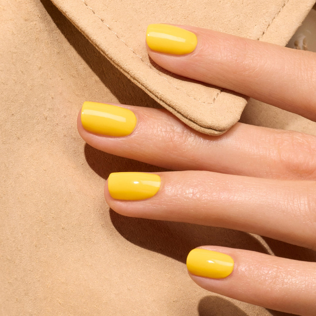 A close-up of a hand with neatly manicured nails painted in Manucurist’s Gold Button gel polish, resting on beige fabric.