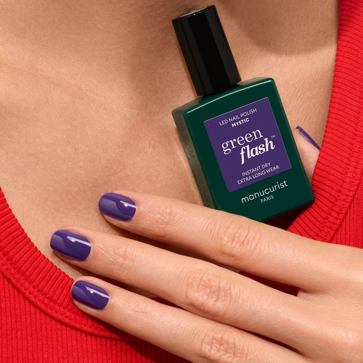 Une main aux ongles peints en violet, vêtue d’un haut rouge, tient un flacon vert de vernis Mystic de Manucurist pour un fini opaque.