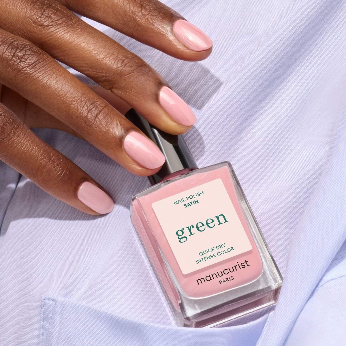 A hand with neatly manicured nails painted in pale pink holds a bottle of Manucurist Satin nail polish, perfectly matching the elegant shade. The person is dressed in a light lavender shirt.