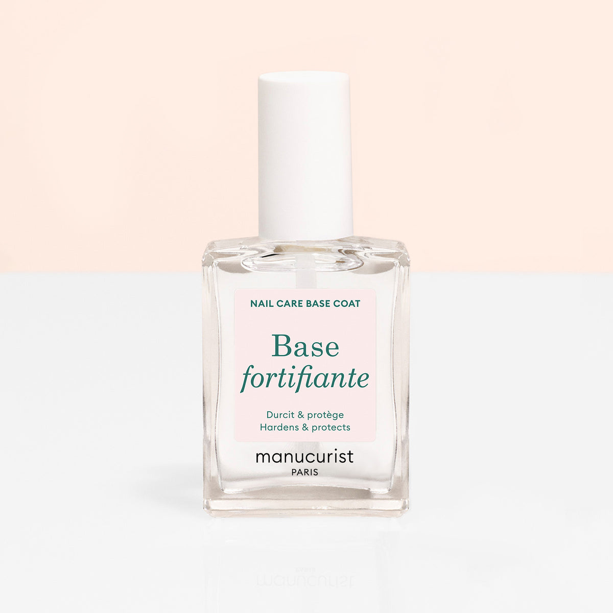 Un flacon en verre transparent du durcisseur Force Manucurist avec un bouchon blanc et une étiquette indiquant "Base fortifiante" et "Hardens protects," sur un fond doux beige et blanc.