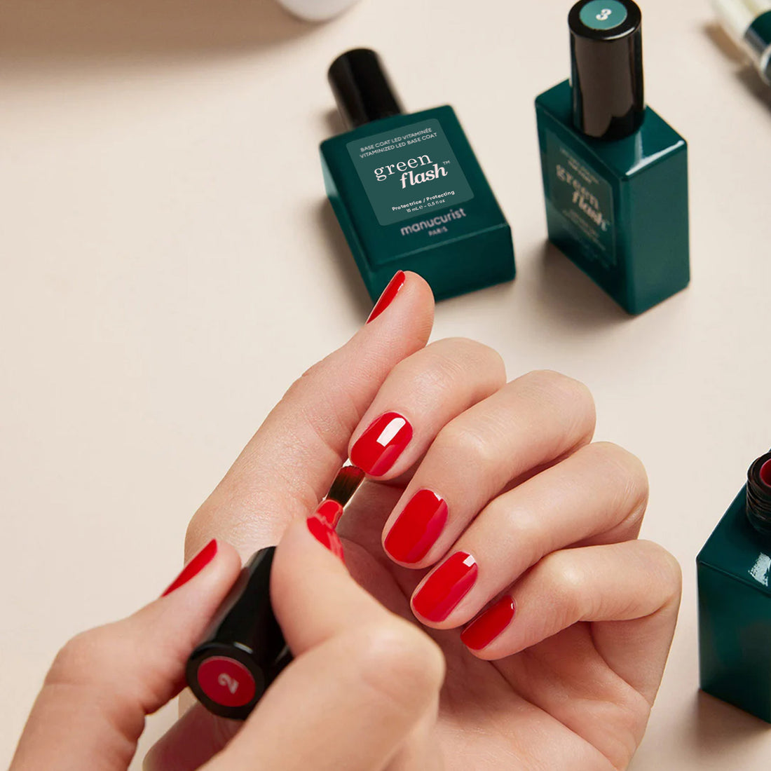 A person applies bright red nail polish as several Manucurist Green Flash™ Base Top Remover Trio bottles and tools are visible on the table in the background.