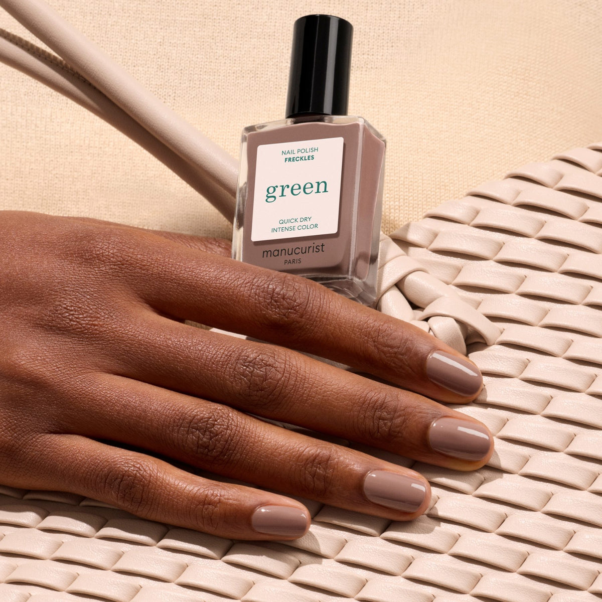Une main manucurée avec le vernis Freckles de Manucurist repose sur un sac en tissu tressé crème, tenant le flacon de vernis pour mettre en valeur un style chic sur un fond beige.
