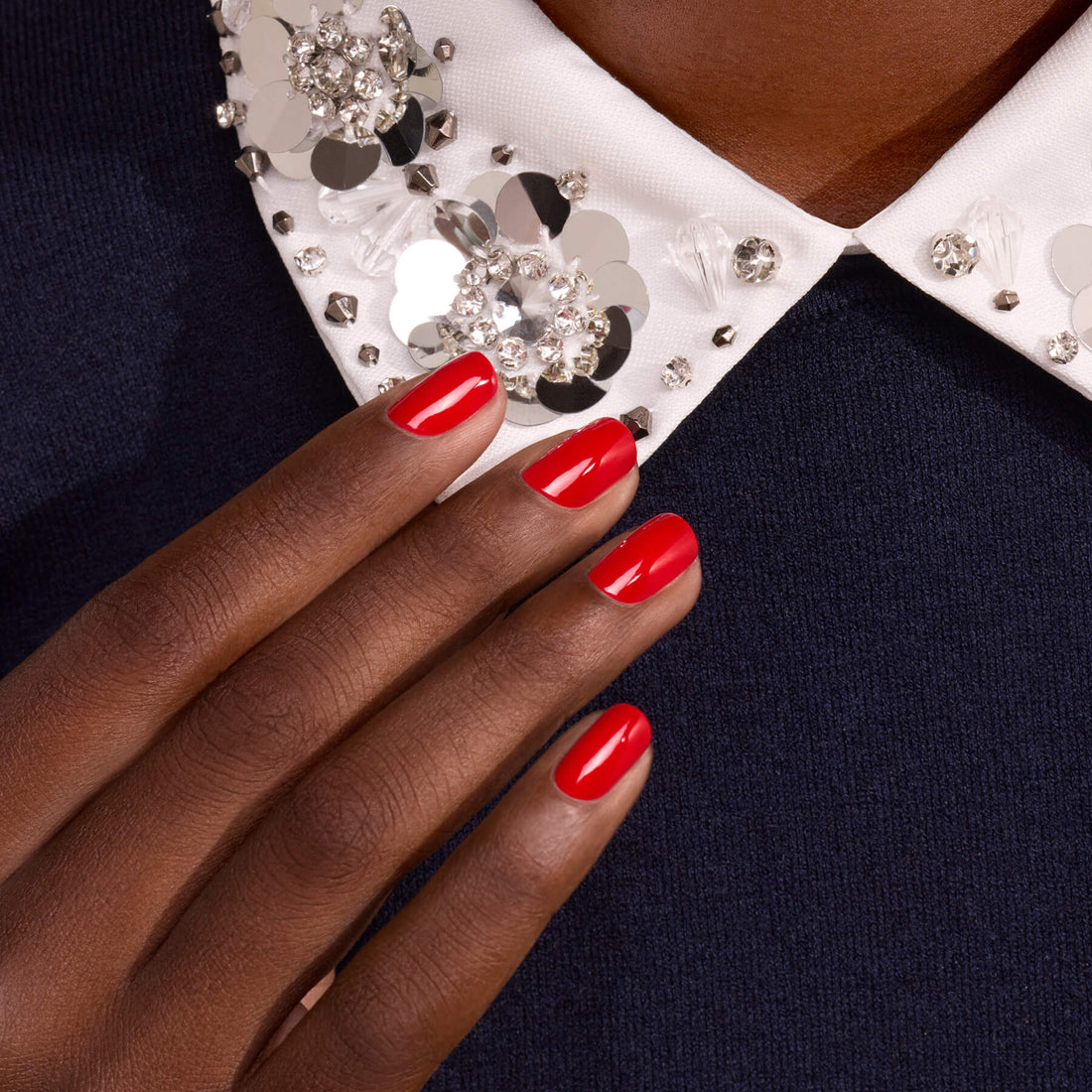 A hand with Anémone by Manucurist—a vivid red polish with blue undertones—rests on dark blue fabric near a white collar adorned with large, sparkling rhinestones and sequins.