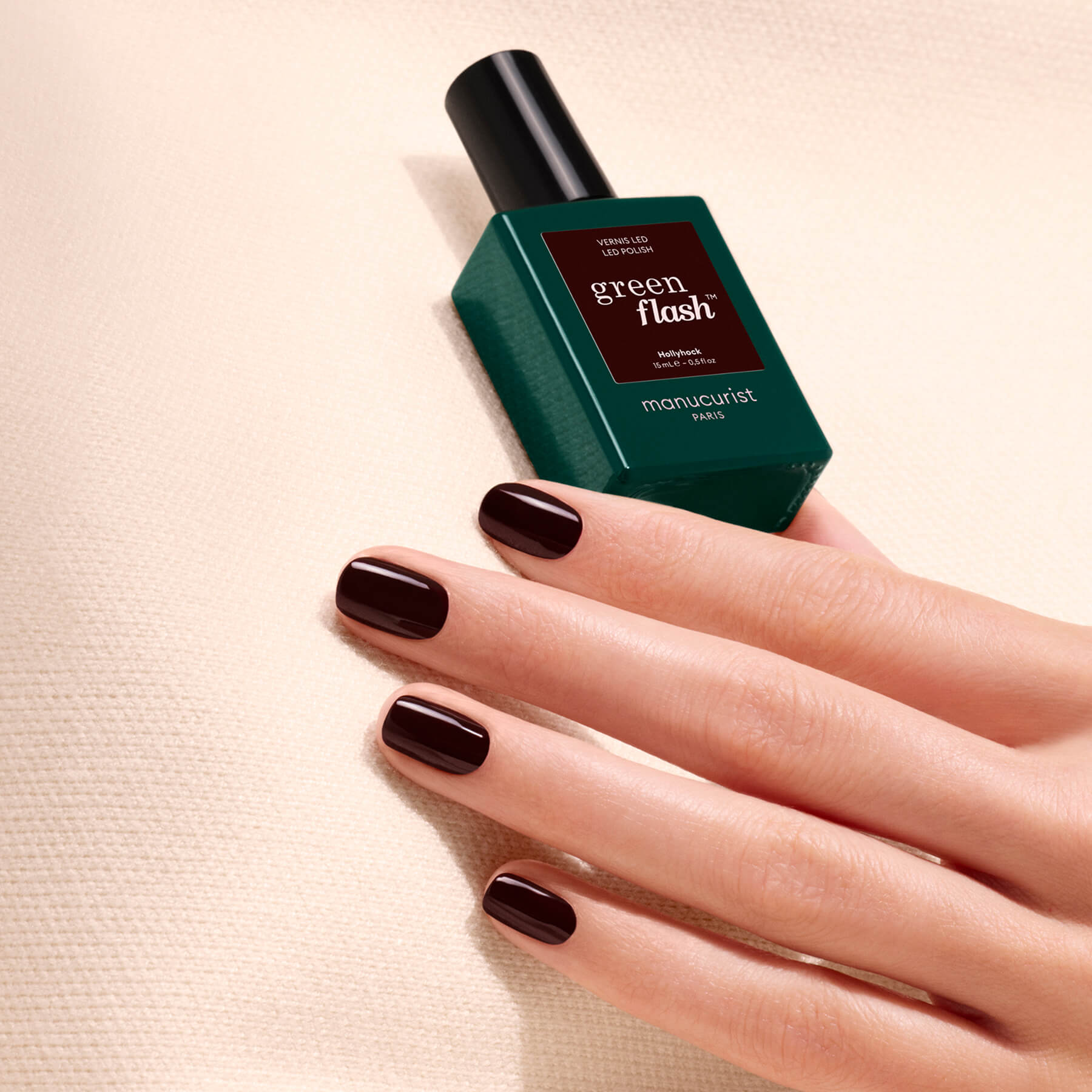 A hand with short, dark burgundy polished nails—reminiscent of a deep red nail polish—rests on a textured cream surface next to a green bottle of Manucurist’s Green Flash gel polish in the shade “Hollyhock.”.