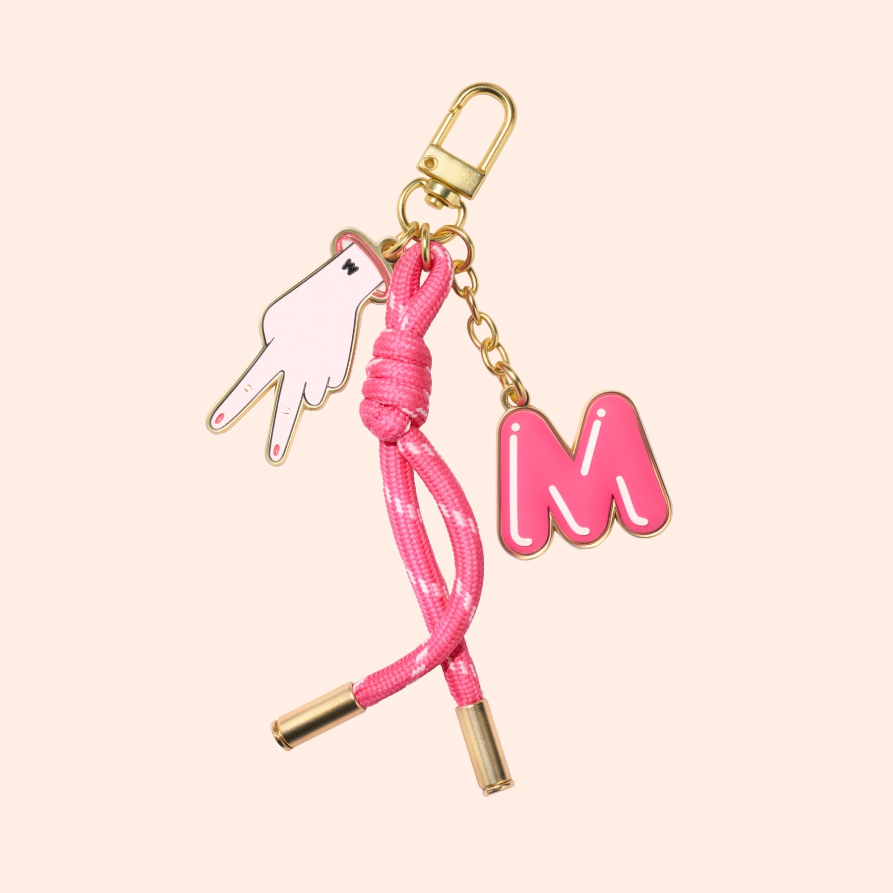 The Manucurist Jelly Charm keychain features a gold clasp, pink and white peace sign hand charm, pink letter M charm, and a pink cord with gold tips—set against a light beige background. Perfect for playful accessory lovers.