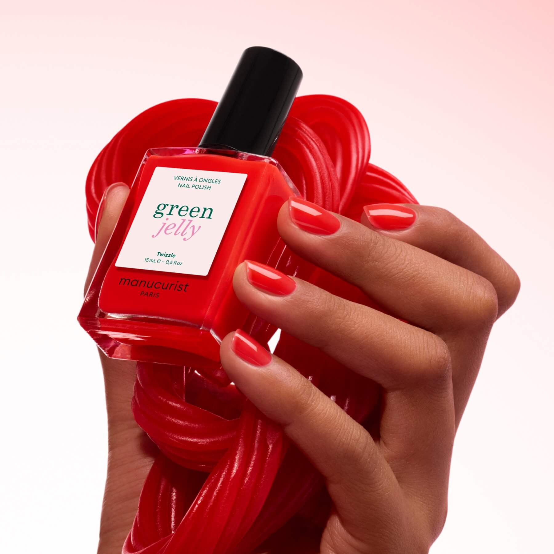 A hand with neatly manicured red nails holds a bottle of Manucurist Twizzle translucent red jelly nail polish against a coil of red licorice, set on a soft pink gradient background.
