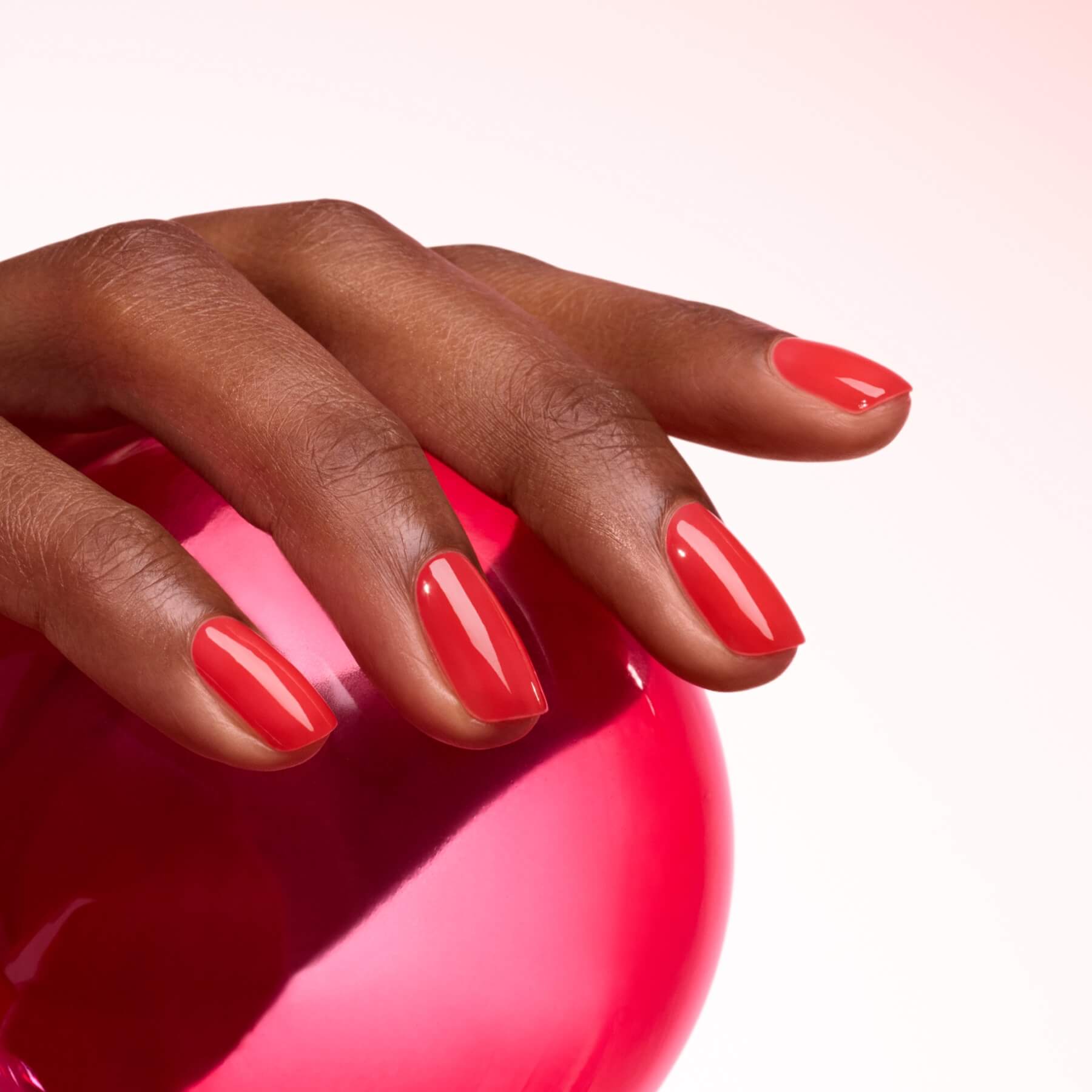 A hand with neatly manicured, ultra-glossy red Twizzle nails by Manucurist rests on a shiny pink sphere against a light pink background.