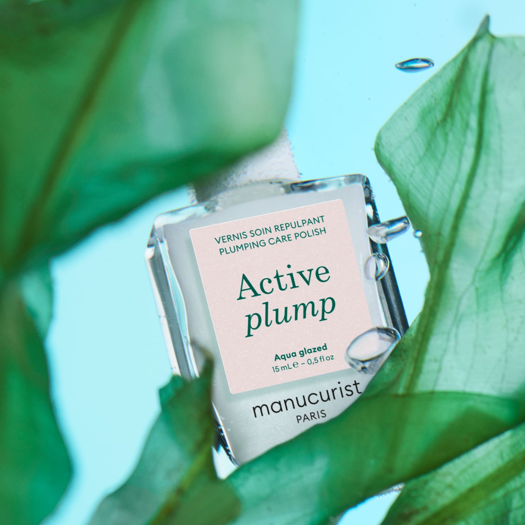 A bottle of Manucurist Active Plump Aqua Glazed nail polish sits among green leaves on a blue background, with water droplets on the bottle.