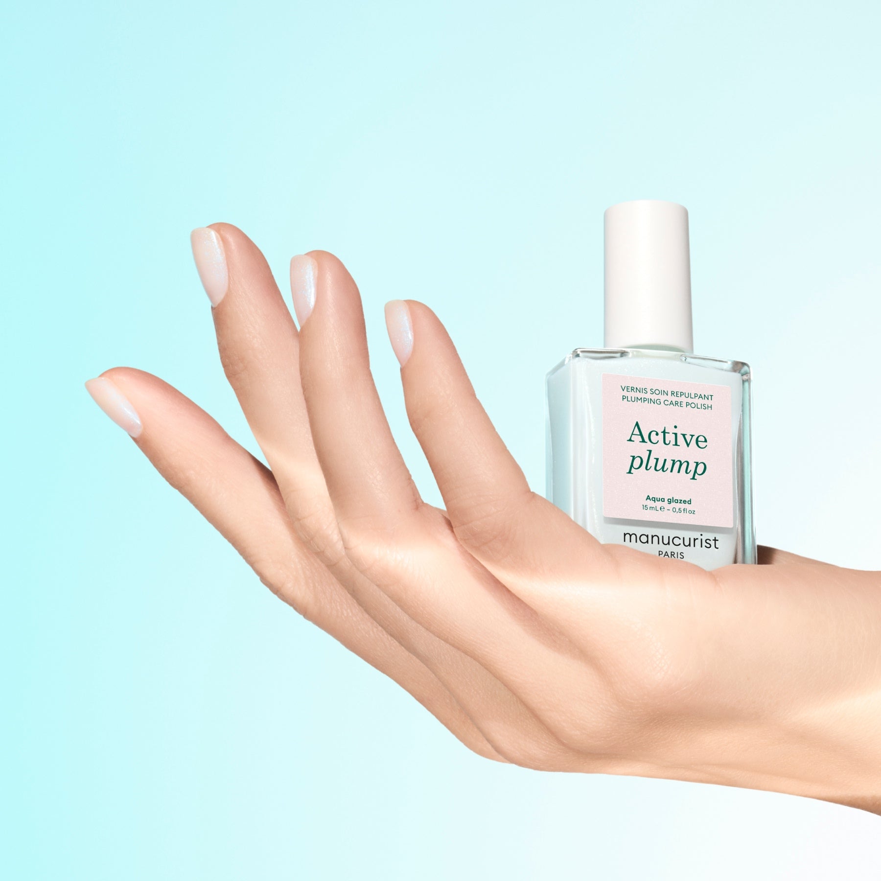 A hand with neatly manicured nails holds a clear bottle of Manucurist Active Plump Aqua Glazed, a nail care product for hydrated nails, against a light blue gradient background.