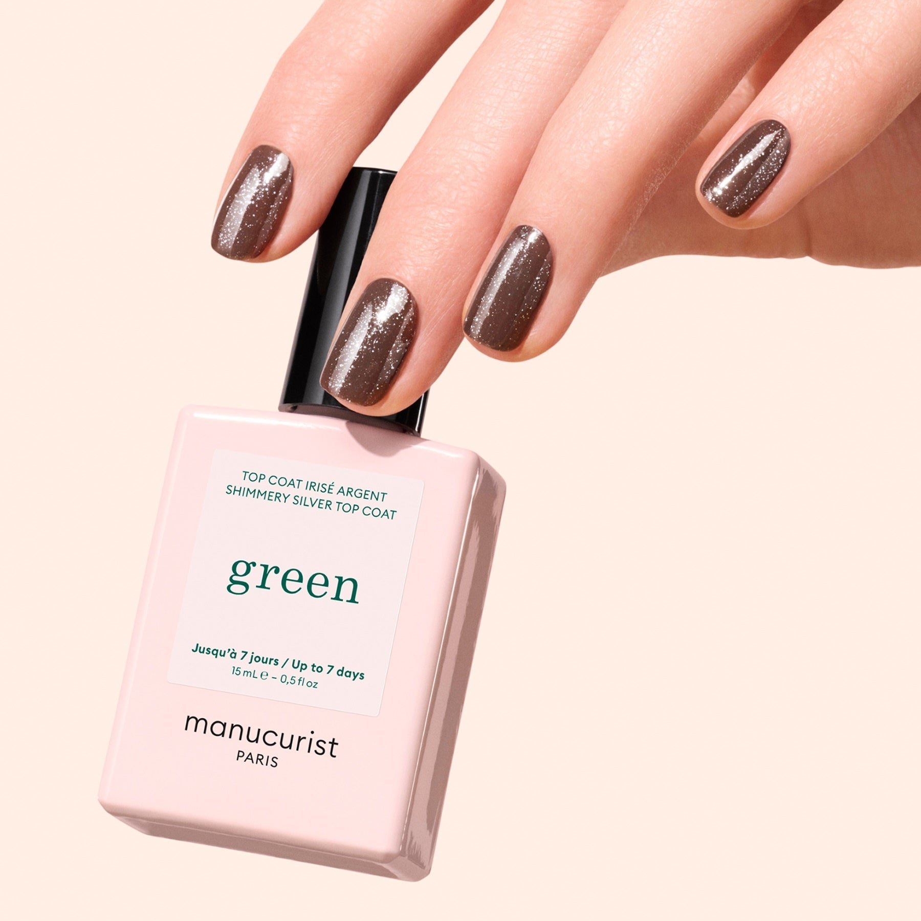 A hand with manicured nails painted in shimmery brown polish holds a bottle of Manucurist Glitter Top Coat, highlighting a dazzling gel-like finish against a light beige background.